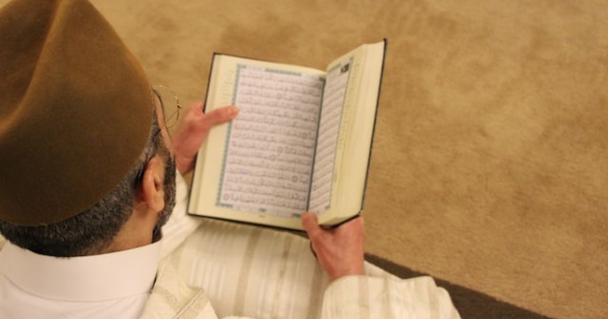 seorang pria muslim duduk dan membaca Al-Quran di tengah karpet berwarna krem